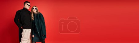 loving young couple in stylish coats with sunglasses posing together on red backdrop, banner