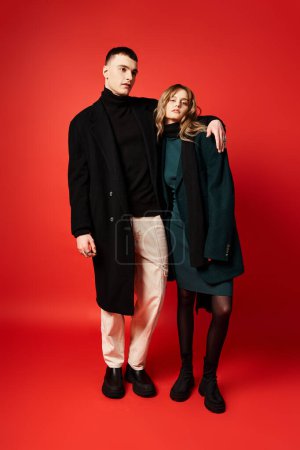 debonair woman in coat looking at camera and posing lovingly with her boyfriend on red backdrop