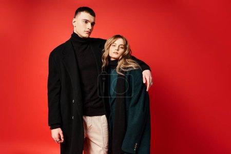 debonair woman in coat looking at camera and posing lovingly with her boyfriend on red backdrop