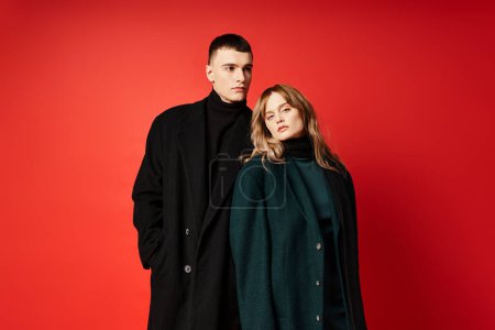 elegant woman in coat looking at camera and posing lovingly with her boyfriend on red backdrop