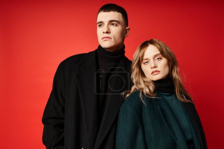 beautiful woman in coat looking at camera and posing lovingly with her boyfriend on red backdrop