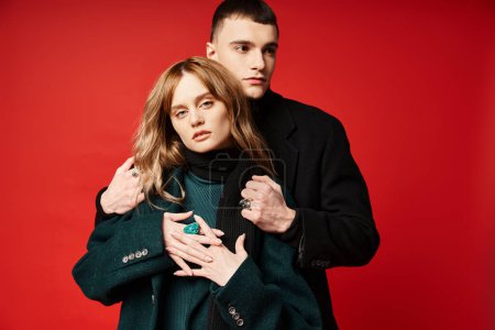 appealing woman in coat looking at camera and posing lovingly with her boyfriend on red backdrop