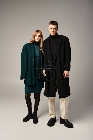 Téléchargez les photos : Couple sophistiqué dans des manteaux élégants posant ensemble sur fond gris et regardant la caméra - en image libre de droit