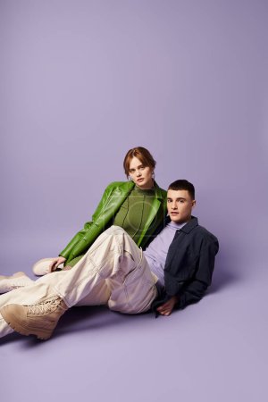well dressed couple in vibrant attires sitting on floor and looking at camera on purple backdrop