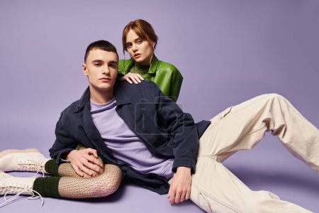 sophisticated couple in vibrant attires sitting on floor and looking at camera on purple backdrop