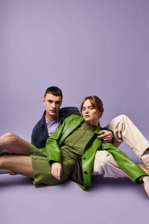 pareja sofisticada en trajes vibrantes sentado en el suelo y mirando a la cámara en el telón de fondo púrpura