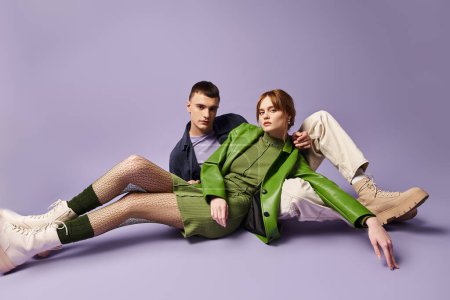 pareja de moda en trajes vibrantes sentado en el suelo y mirando a la cámara en el telón de fondo púrpura