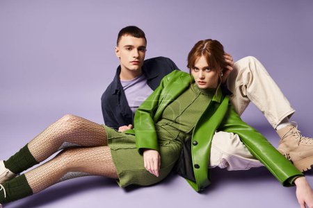 Téléchargez les photos : Couple à la mode en vêtements vibrants assis sur le sol et en regardant la caméra sur fond violet - en image libre de droit