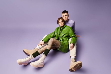 appealing couple in vibrant attires sitting on floor and looking at camera on purple backdrop