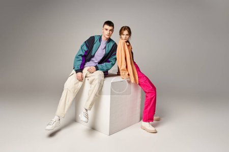loving fashionable couple in vivid stylish bombers looking at camera on gray backdrop on white cube