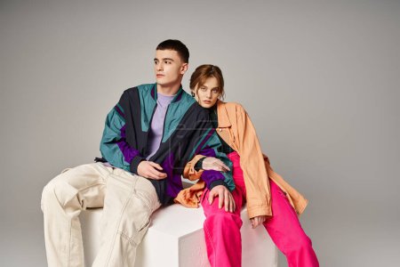 alluring woman in fashionable bomber looking at camera next to her handsome boyfriend on white cube