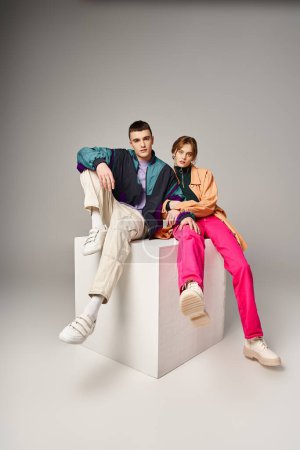 caring fashionable couple in vivid stylish bombers looking at camera on gray backdrop on white cube