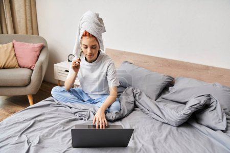 ansprechende rothaarige queere Person in Hausbekleidung mit Gesichtsroller, während sie es sich mit Laptop im Bett gemütlich macht