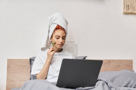 atractivo pelirrojo queer persona en ropa de casa usando rodillo facial mientras se relaja en la cama con el ordenador portátil