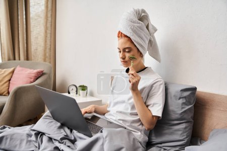 Foto de Atractivo pelirrojo queer persona en ropa de casa utilizando rodillo facial mientras se relaja en la cama con el ordenador portátil - Imagen libre de derechos