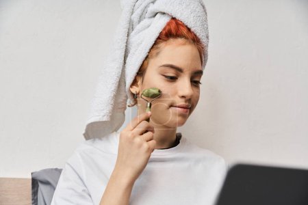 attractive red haired queer person in homewear using face roller while relaxing on bed with laptop