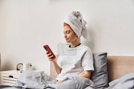 Photo for Young extravagant person in homewear with hair towel looking at her smartphone while relaxing in bed - Royalty Free Image