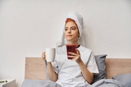 Foto de Persona queer relajante positivo en ropa de casa con toalla de pelo usando el teléfono y beber té en la cama - Imagen libre de derechos
