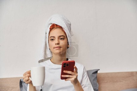 Téléchargez les photos : Positif relaxant queer personne dans les vêtements de maison avec serviette de toilette en utilisant le téléphone et boire du thé au lit - en image libre de droit