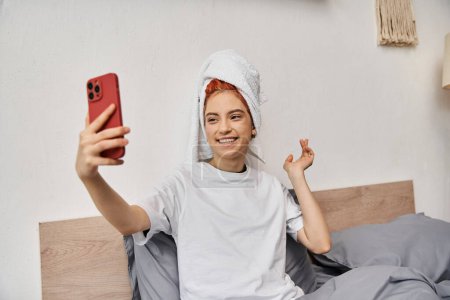 Foto de Alegre hermosa persona queer con toalla de pelo en la ropa de casa tomar selfies mientras se relaja en la cama - Imagen libre de derechos