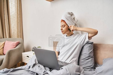 Téléchargez les photos : Joyeuse jolie personne queer avec serviette de cheveux regarder des films sur ordinateur portable et en utilisant gua sha tout au lit - en image libre de droit