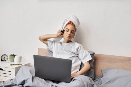 Foto de Alegre bastante queer persona con toalla de pelo viendo películas en el ordenador portátil y el uso de gua sha mientras está en la cama - Imagen libre de derechos