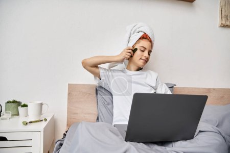 alegre bastante queer persona con toalla de pelo viendo películas en el ordenador portátil y utilizando gua sha en la cama
