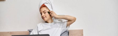 joyeux jolie personne queer avec serviette de cheveux regarder des films sur ordinateur portable et en utilisant gua sha au lit, bannière