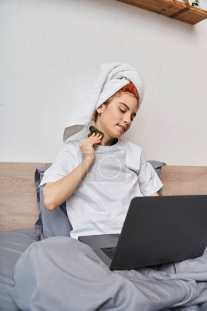 Foto de Alegre bastante queer persona con toalla de pelo viendo películas en el ordenador portátil y utilizando gua sha en la cama - Imagen libre de derechos