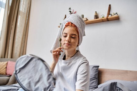 Photo for Good looking queer person in cozy homewear using face roller in front of mirror as morning routine - Royalty Free Image
