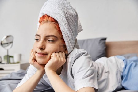 fröhliche schöne queere Person in Hauskleidung mit Haartuch, die in ihrem Bett chillt und wegschaut