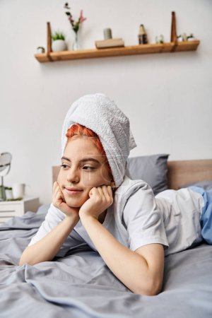 fröhliche schöne queere Person in Hauskleidung mit Haartuch, die in ihrem Bett chillt und wegschaut