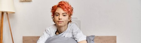 Foto de Somnoliento extravagante queer persona en ropa de casa con el pelo rojo sentado en la cama mirando a la cámara, pancarta - Imagen libre de derechos