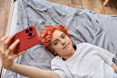 junge entspannte queere Person in Hauskleidung mit roten Haaren, die auf dem Bett liegt und Selfies mit ihrem Handy macht