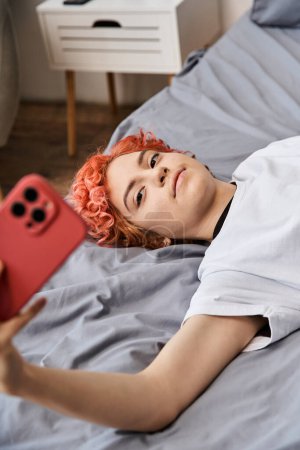 joven relajante queer persona en ropa de casa con el pelo rojo acostado en la cama y tomar selfies en su teléfono