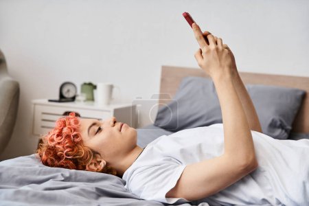 Téléchargez les photos : Séduisante personne queer relaxant dans les vêtements de maison avec les cheveux roux couché sur le lit et en utilisant son smartphone - en image libre de droit