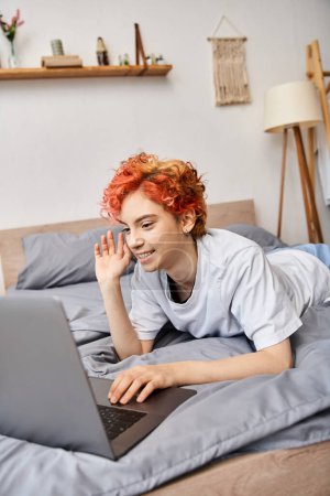 gut aussehende rothaarige fröhliche queere Person in Hauskleidung mit Videoanruf, während sie im Bett liegt