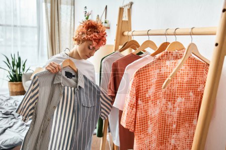 Photo for Beautiful red haired extravagant person picking up stylish clothes near rack at home, leisure time - Royalty Free Image