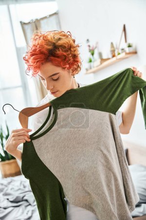 appealing red haired extravagant person picking up stylish clothes near rack at home, leisure time