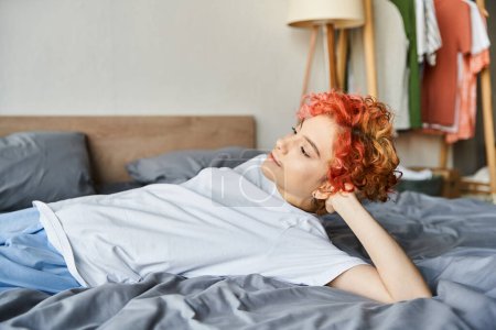 beautiful extravagant person with red vibrant hair lying in her bed and looking away, leisure time