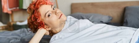 Foto de Atractivo queer persona con el pelo rojo acostado en su cama y mirando a la cámara, tiempo libre, pancarta - Imagen libre de derechos