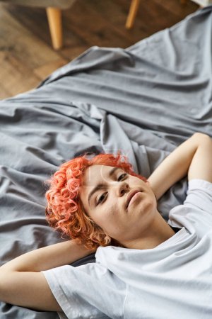 cheerful appealing queer person with red hair lying on her bed and looking at camera, leisure time