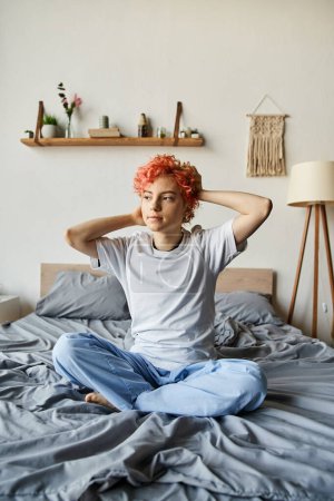 appealing queer person with vibrant red hair sitting on bed and looking away, leisure time