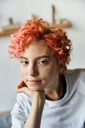 Foto de Hermosa persona queer con el pelo rojo vibrante sentado en la cama y mirando a la cámara, tiempo libre - Imagen libre de derechos