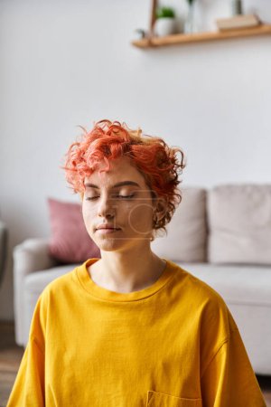 Foto de Hermosa persona queer extravagante con el pelo rojo en vibrante camiseta amarilla meditando en casa - Imagen libre de derechos