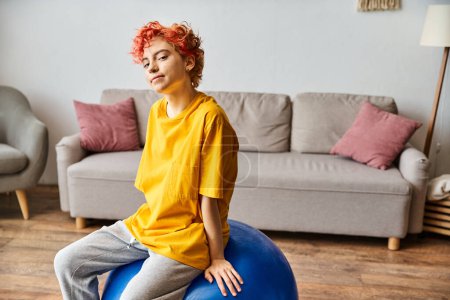 Photo for Cheerful queer person with red hair sitting on fitness ball and smiling at camera while at home - Royalty Free Image