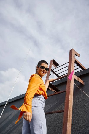 freudige junge kaukasische Frau in lebendiger urbaner Kleidung posiert auf einer Treppe auf dem Dach und lächelt in die Kamera