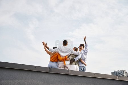 Rückansicht einer Gruppe junger multikultureller Freunde in lässiger urbaner Kleidung, die sich auf dem Dach umarmen
