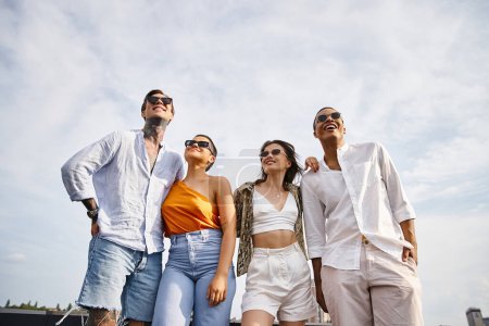 vier verschiedene attraktive, gut gelaunte Freunde mit stylischer Sonnenbrille posieren fröhlich gemeinsam auf dem Dach