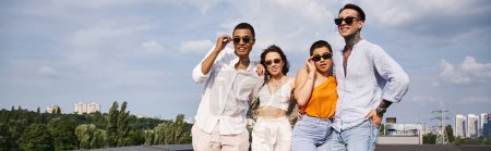 group of four joyous interracial friends with stylish sunglasses posing together on rooftop, banner